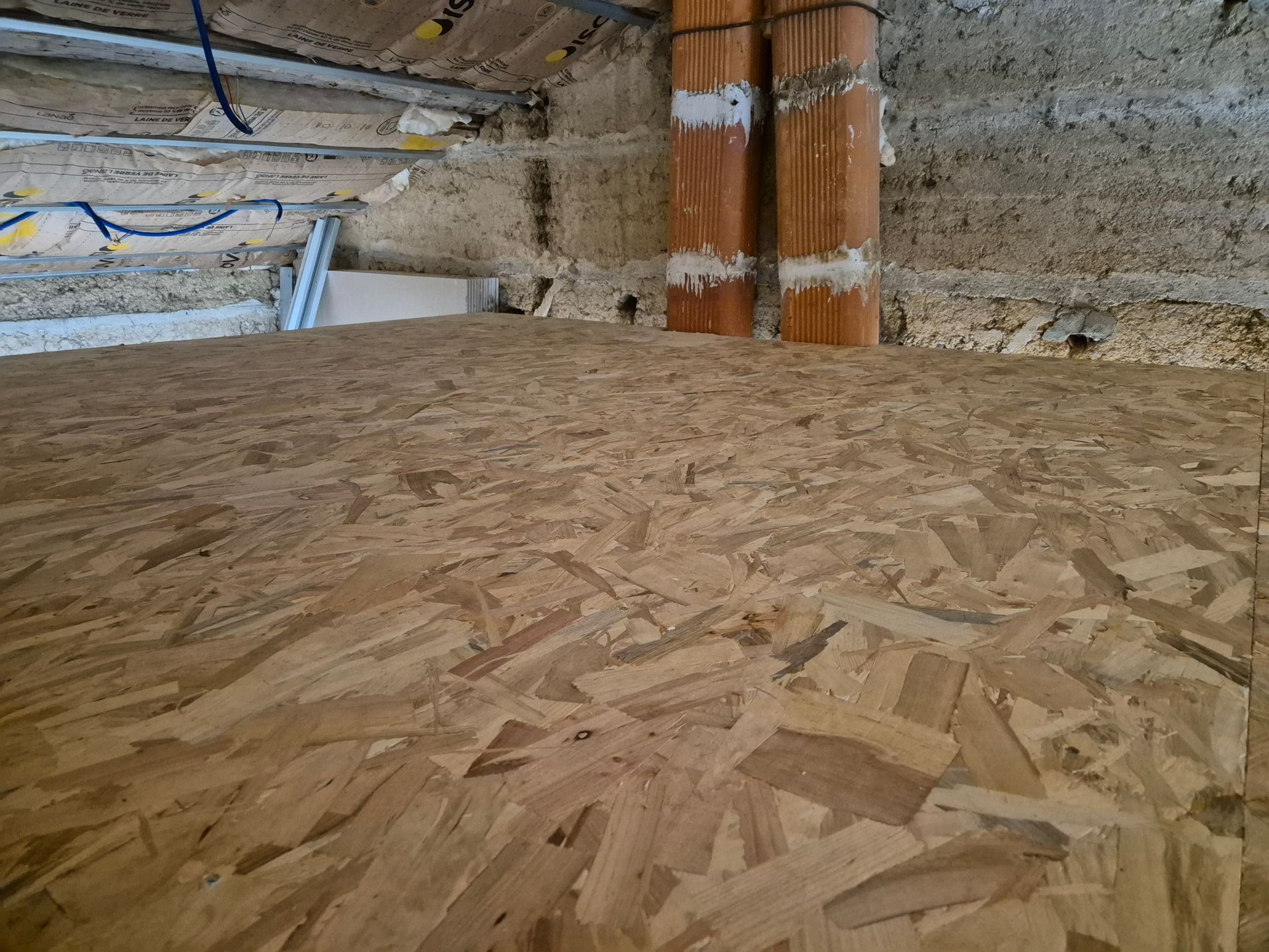 Gros plan sur un sous-plancher en OSB dans un grenier en construction, avec isolation et tuyaux