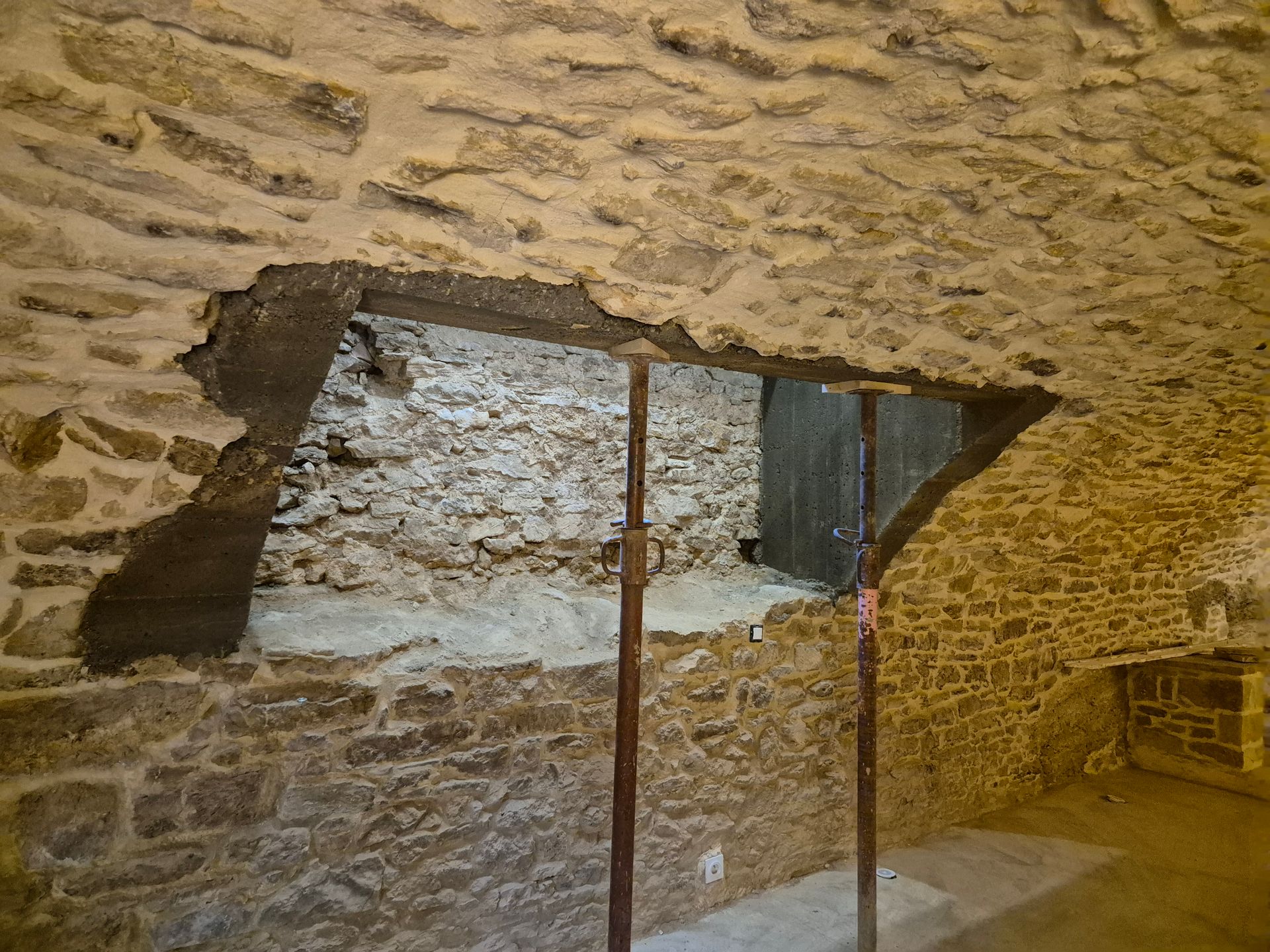 Mur intérieur en pierre avec une petite ouverture rectangulaire et deux barres métalliques rouillées.