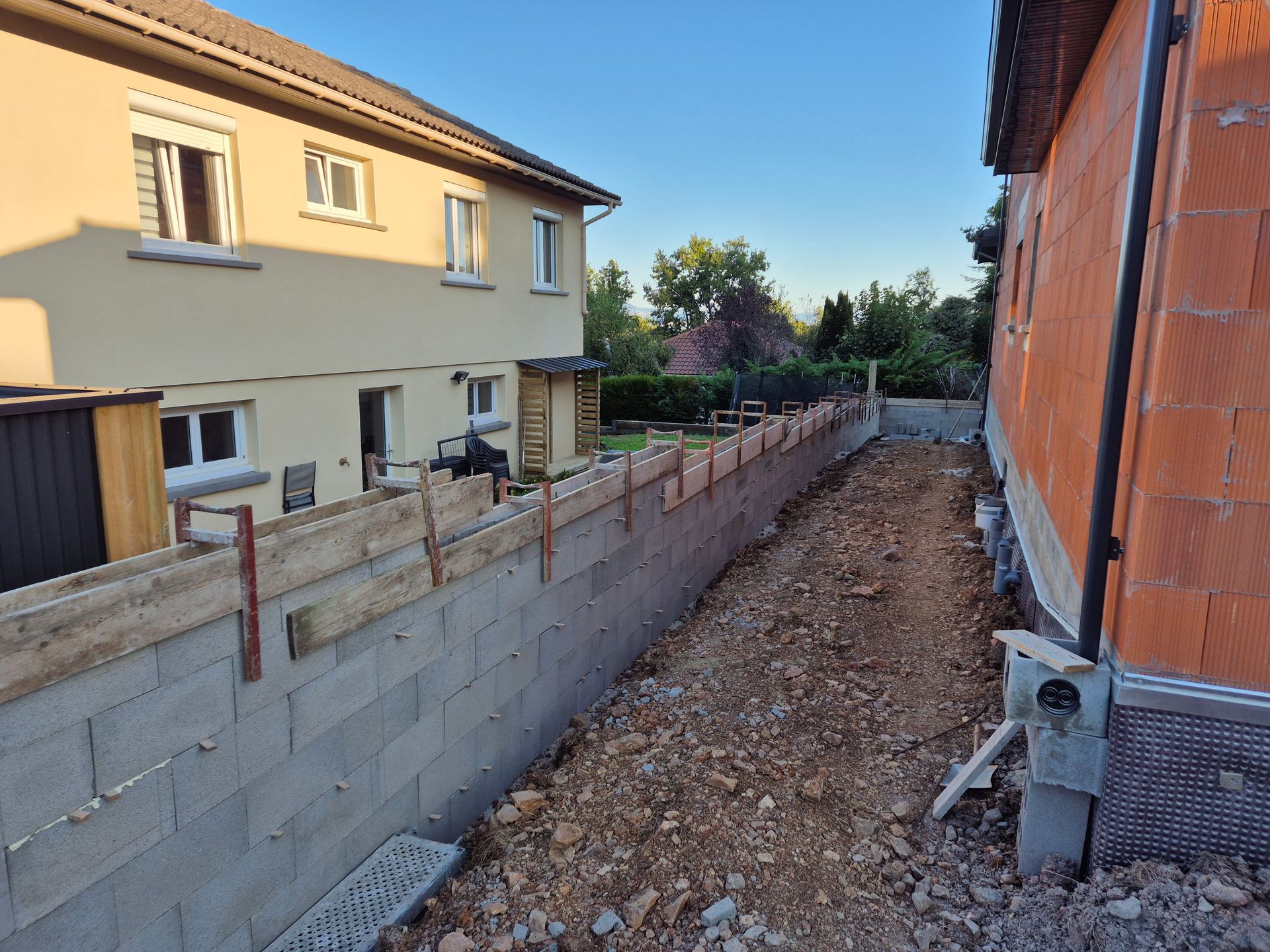 Tranchée de construction étroite entre les maisons, avec un mur en parpaings et des tuyaux apparents