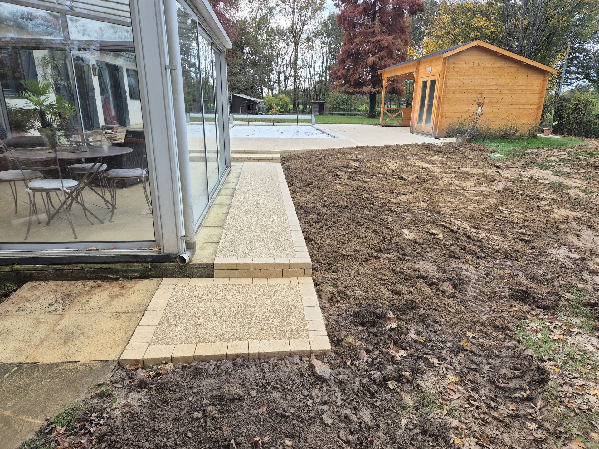 Terrasse arrière avec une véranda, une allée en pierre, une pelouse boueuse et un petit abri en bois.