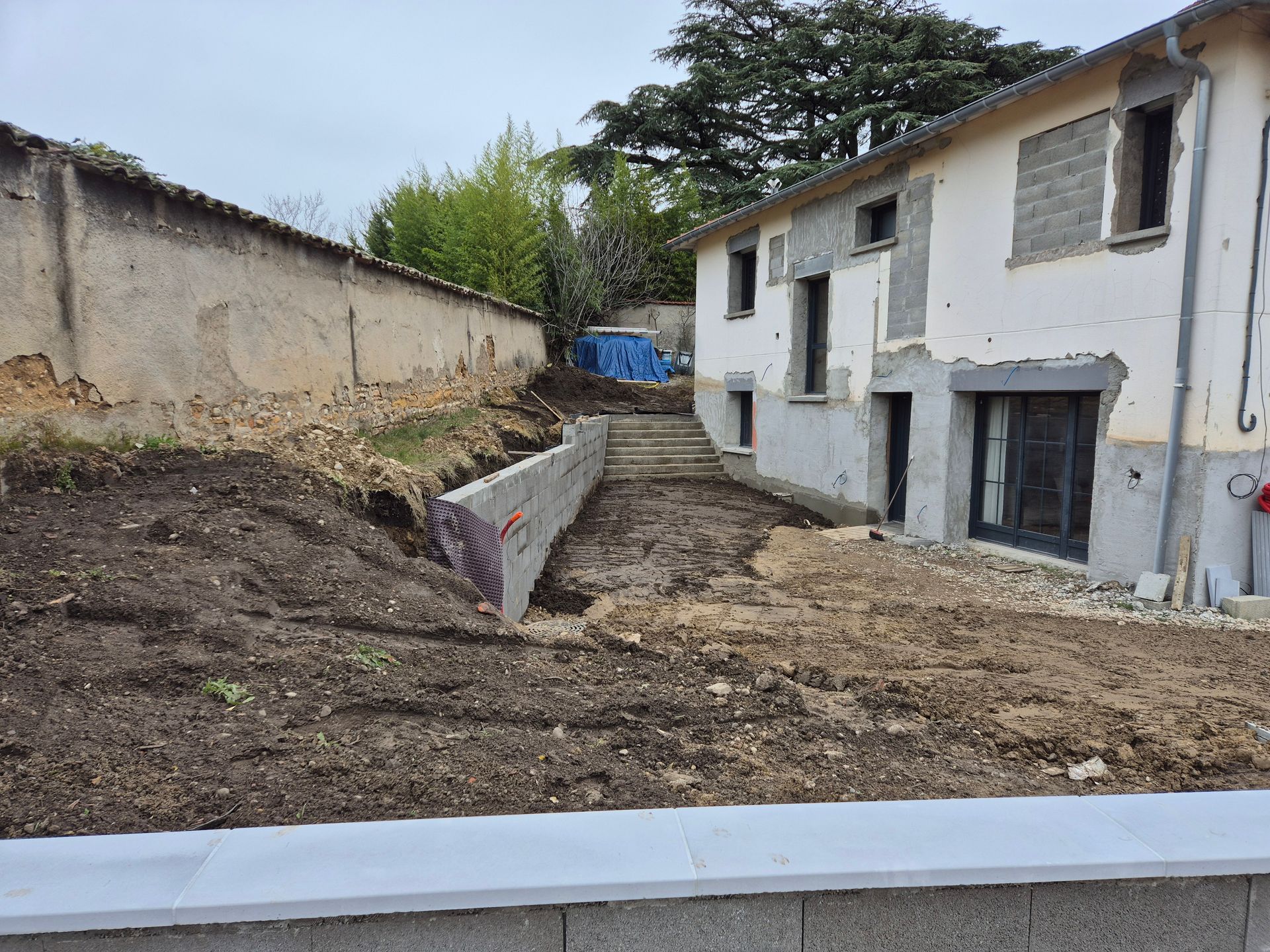 Chantier de construction jouxtant un bâtiment blanc, avec une cour boueuse excavée et un mur de soutènement.