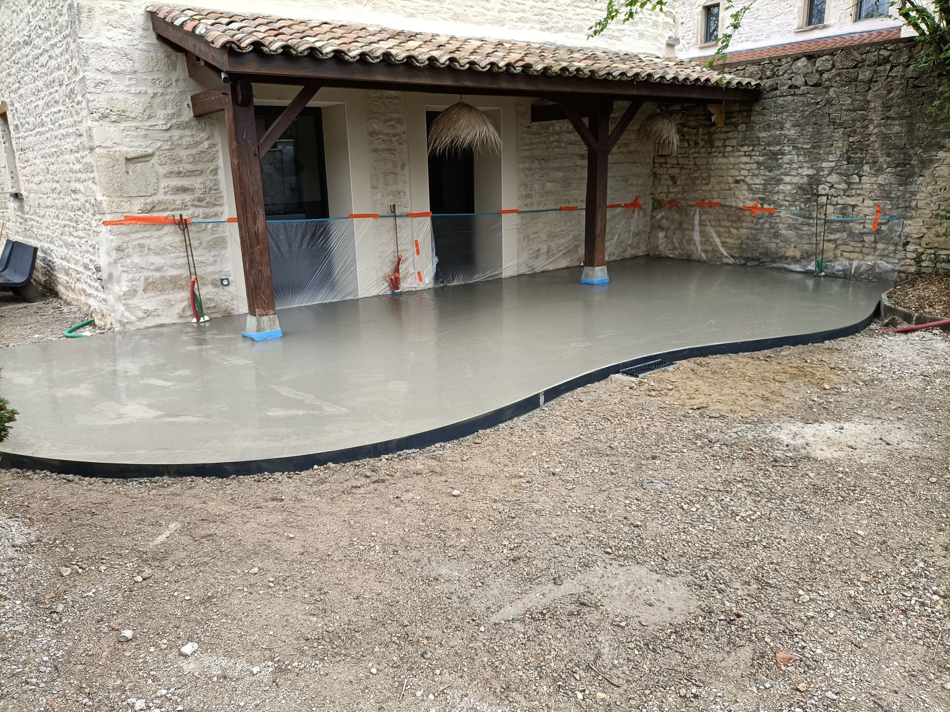 Cour avec dalle juste coulée, un mur de pierre et un porche couvert avec des poteaux sur des supports bleus.