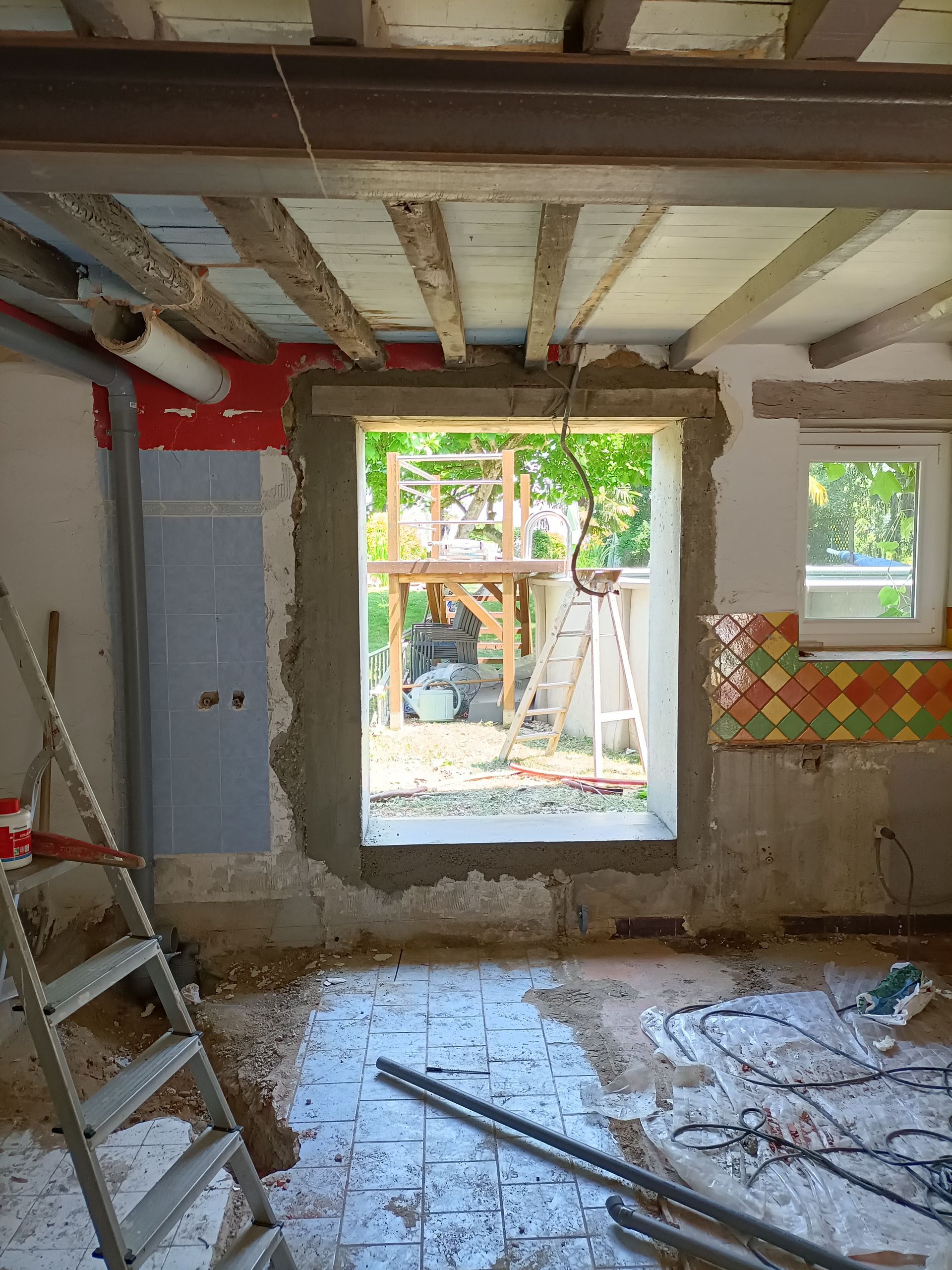 Pièce partiellement rénovée avec poutres apparentes, porte ouverte, échelle et débris de construction au sol