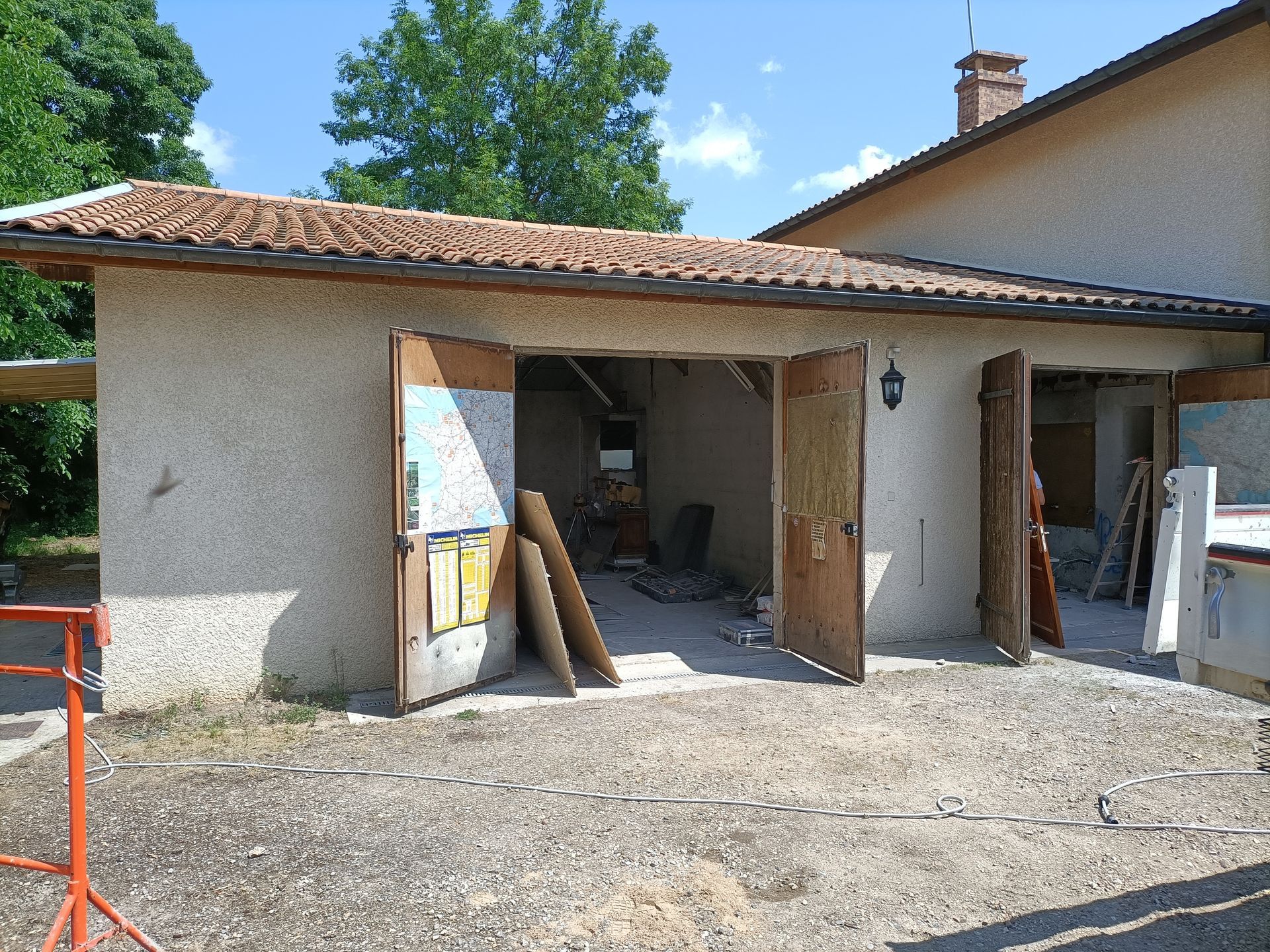 Petit garage avec portes ouvertes, planches de bois et cour de gravier sous un ciel ensoleillé