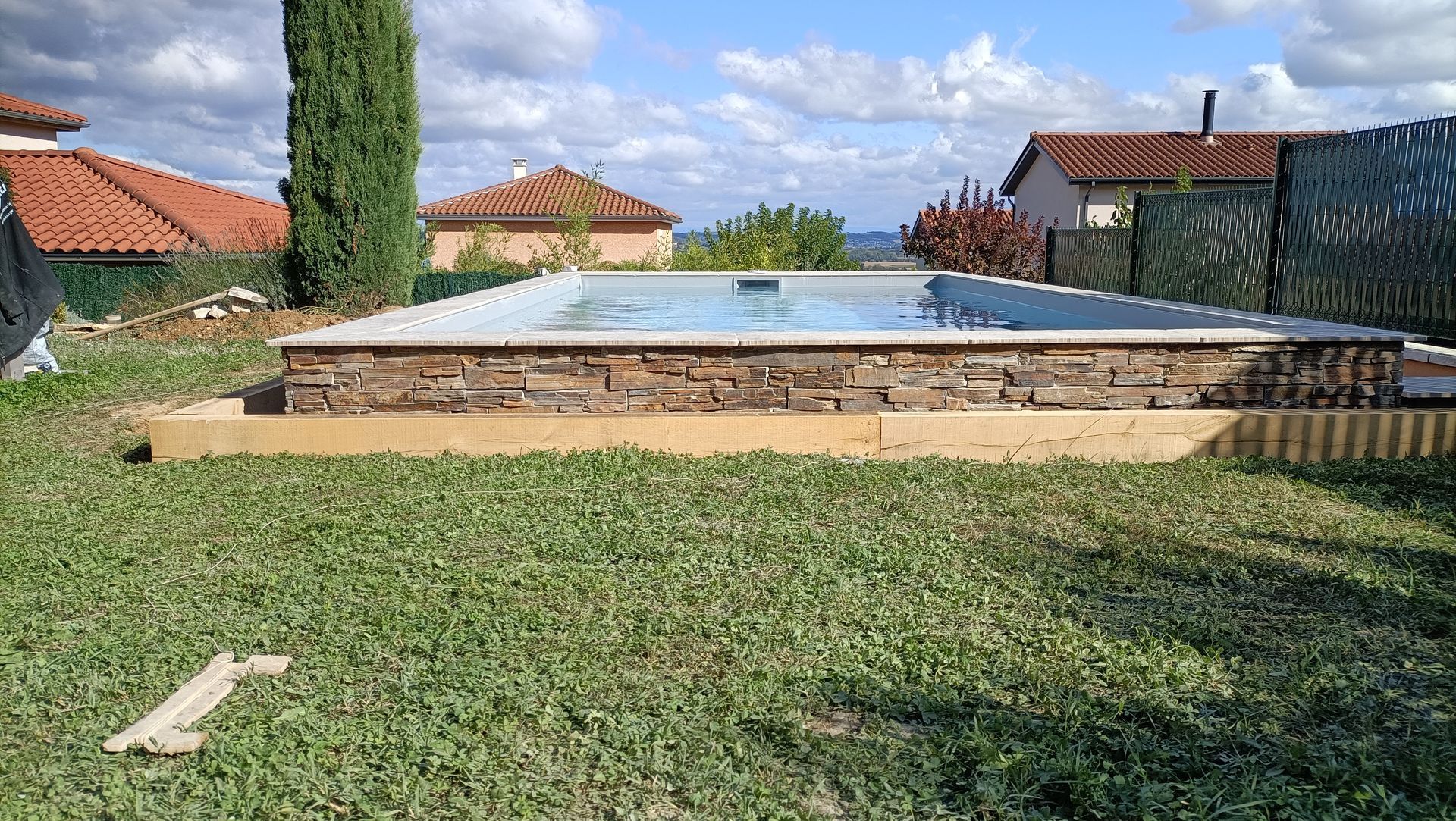 Piscine semi-enterrée à côté d'une pelouse et de maisons aux toits de tuiles sous un ciel partiellement nuageux