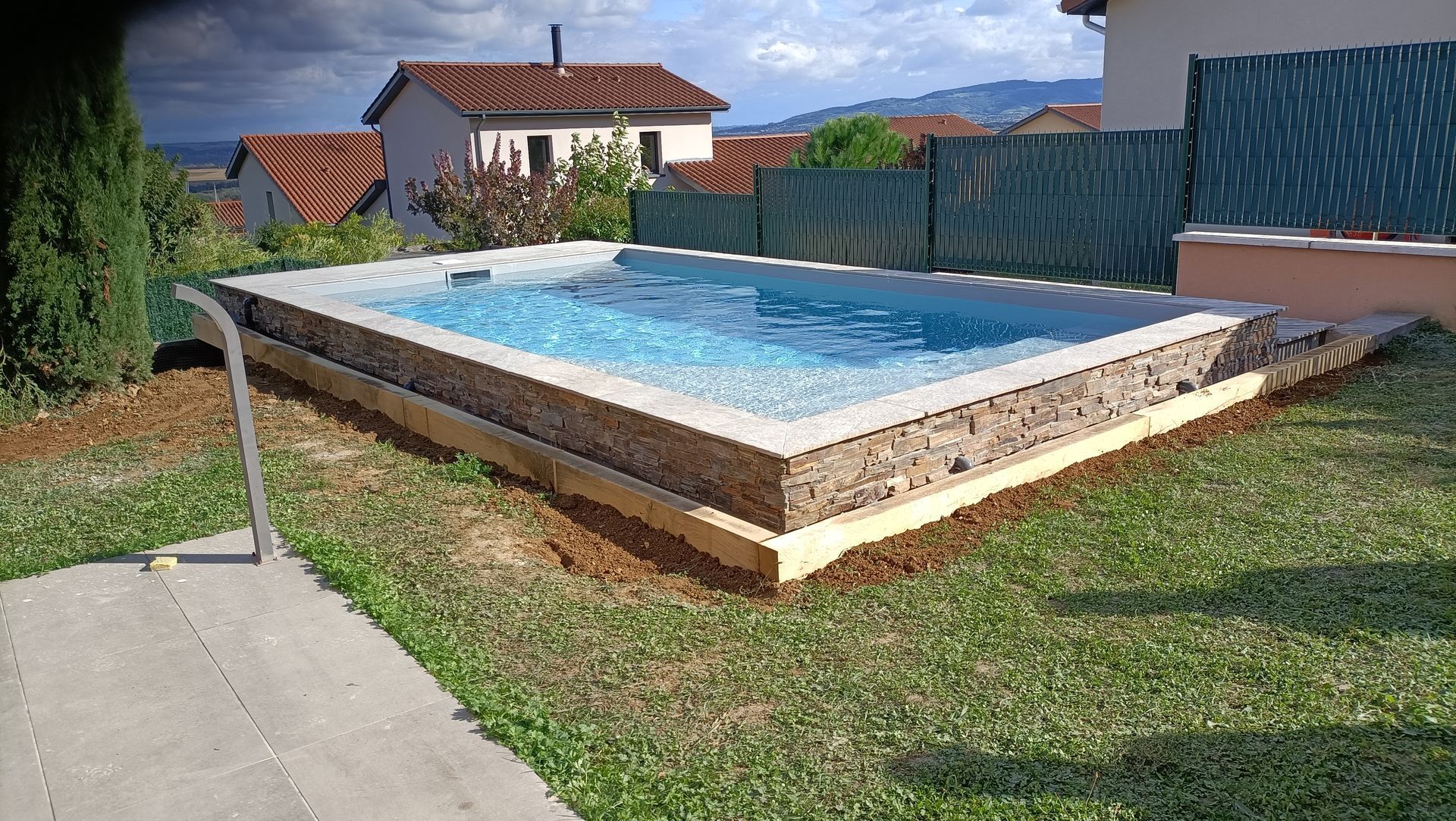 Piscine surélevée dans un jardin à côté d'une pelouse et d'une terrasse, avec des maisons et des collines en arrière-plan.