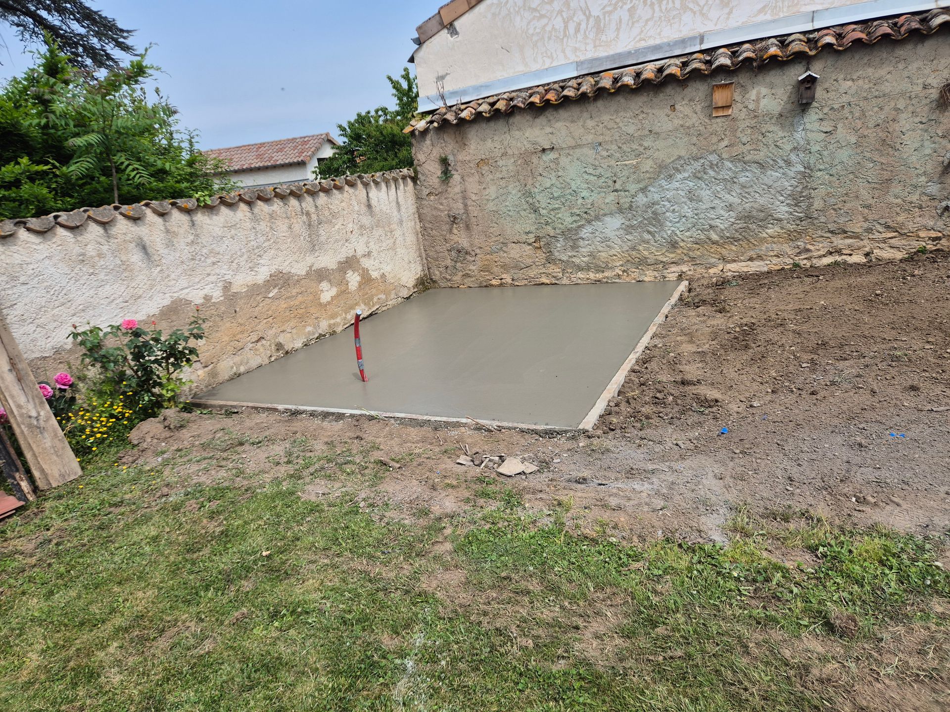 Cour arrière avec une dalle de béton fraîchement coulée à côté d'un mur de pierre et d'une cour en terre.