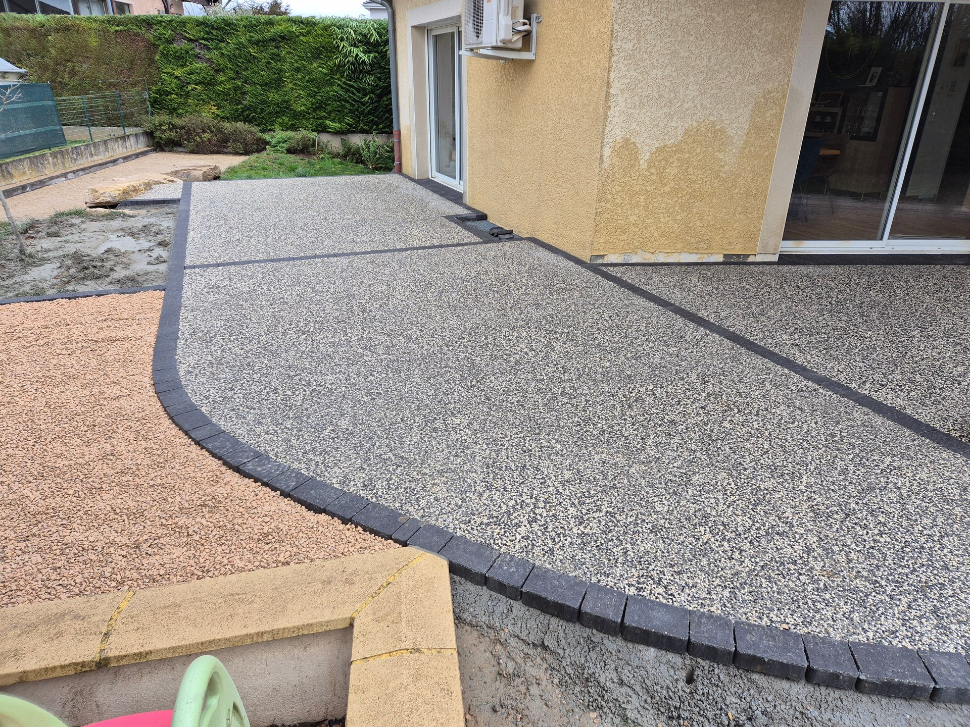 Dallage avec graviers gris et beiges le long d'une maison et porte-fenêtre donnant sur un patio, dans un jardin.