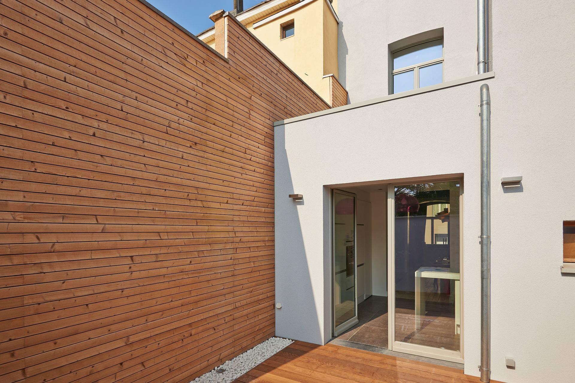 Cour intérieure moderne avec bâtiment blanc, mur revêtu de bois et porte vitrée baignée de soleil