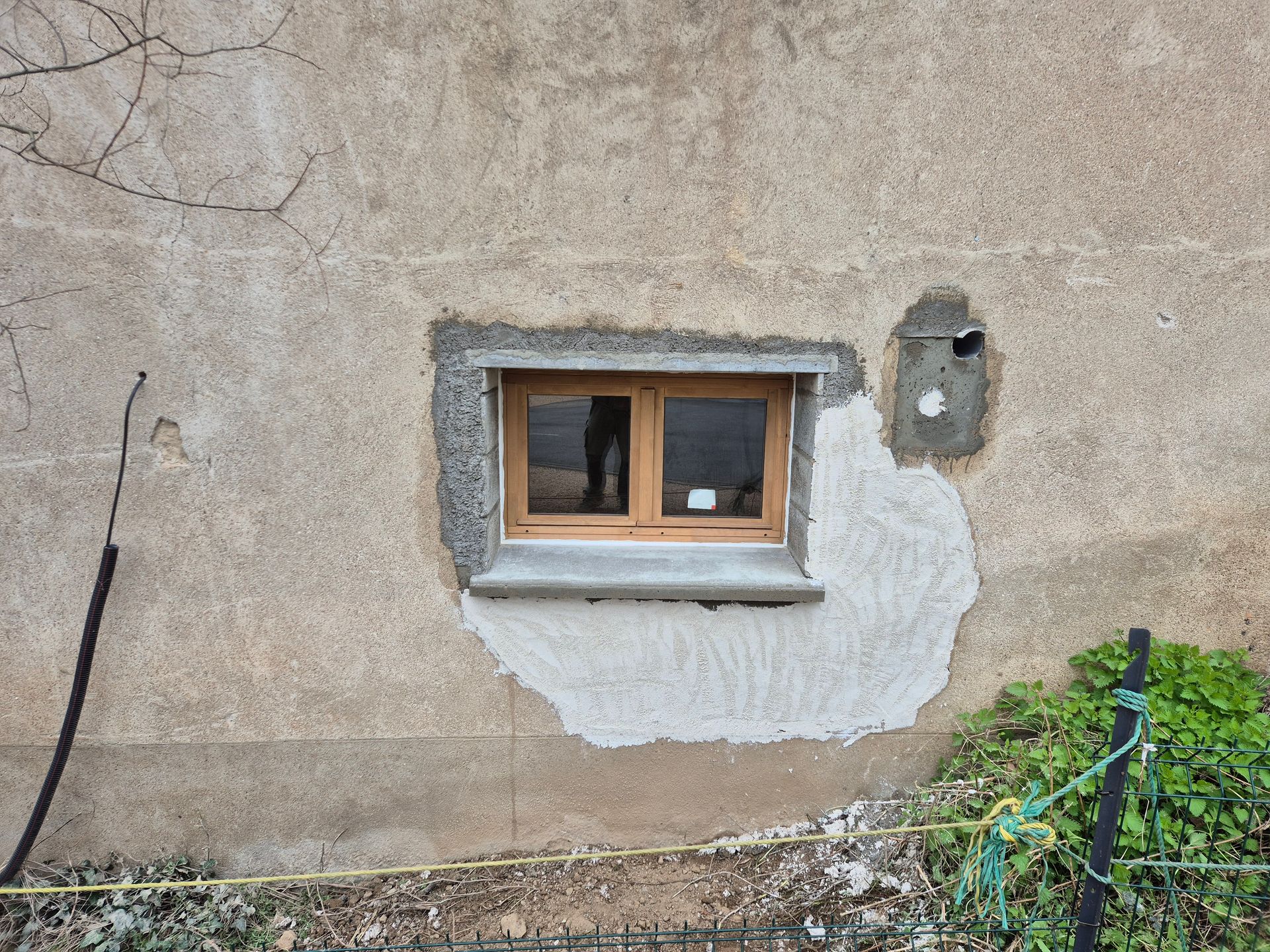 Petite fenêtre dans un mur en stuc défraîchi, avec des réparations blanches et des vignes en bas à droite