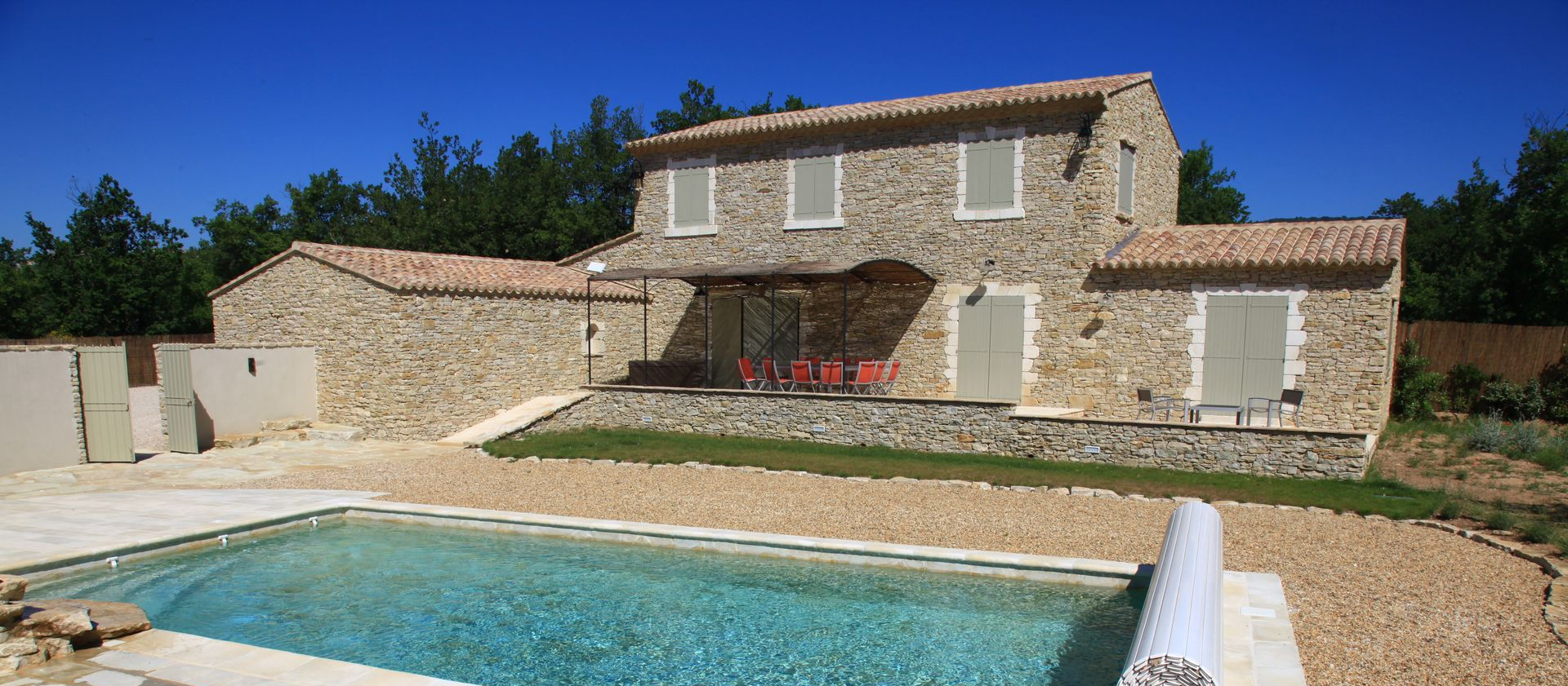 Maison en pierre au toit de tuiles rouges, jouxtant une piscine dans une cour ensoleillée.