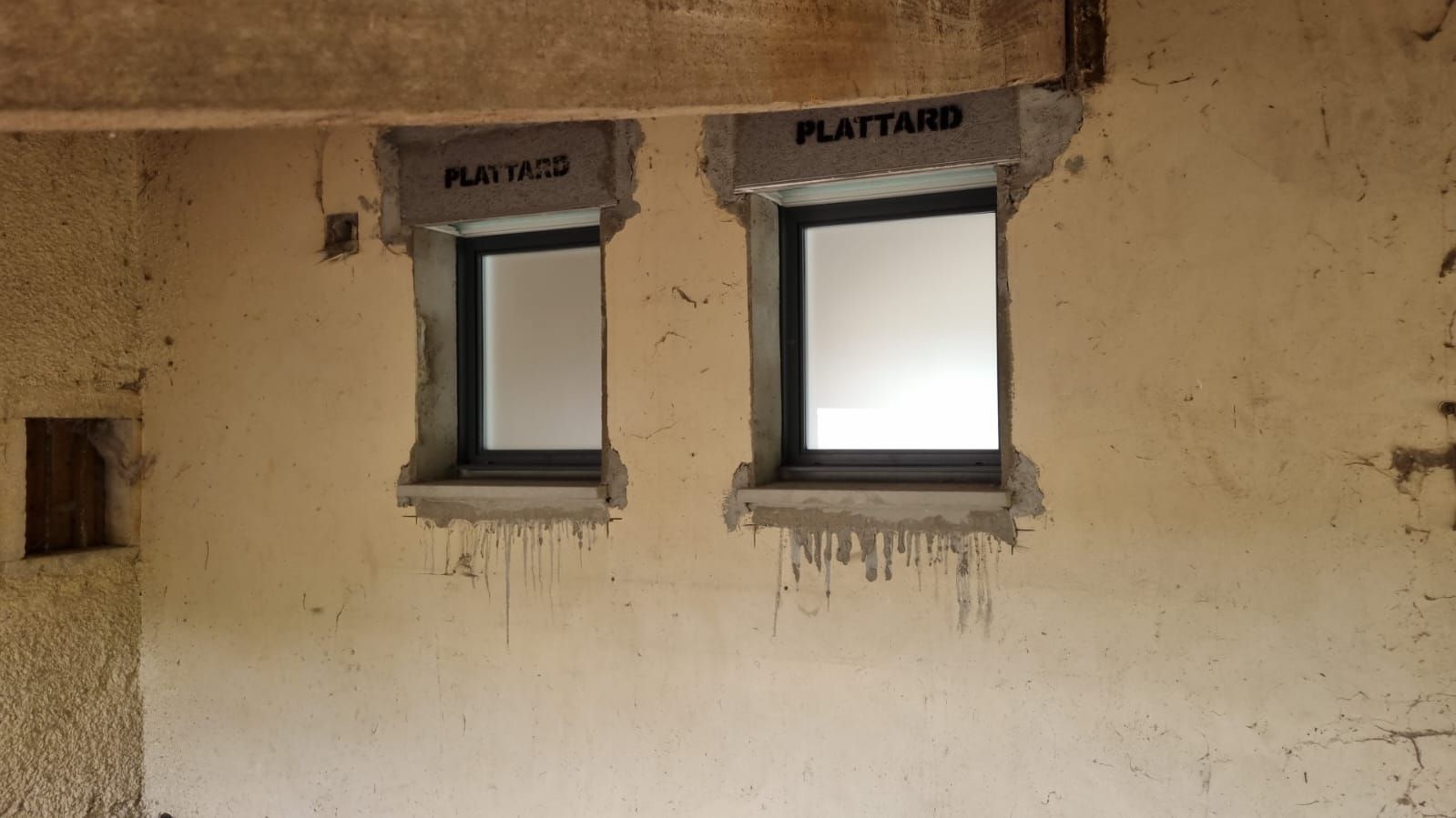 Deux petites fenêtres à barreaux dans un mur beige rugueux, avec des inscriptions au-dessus.