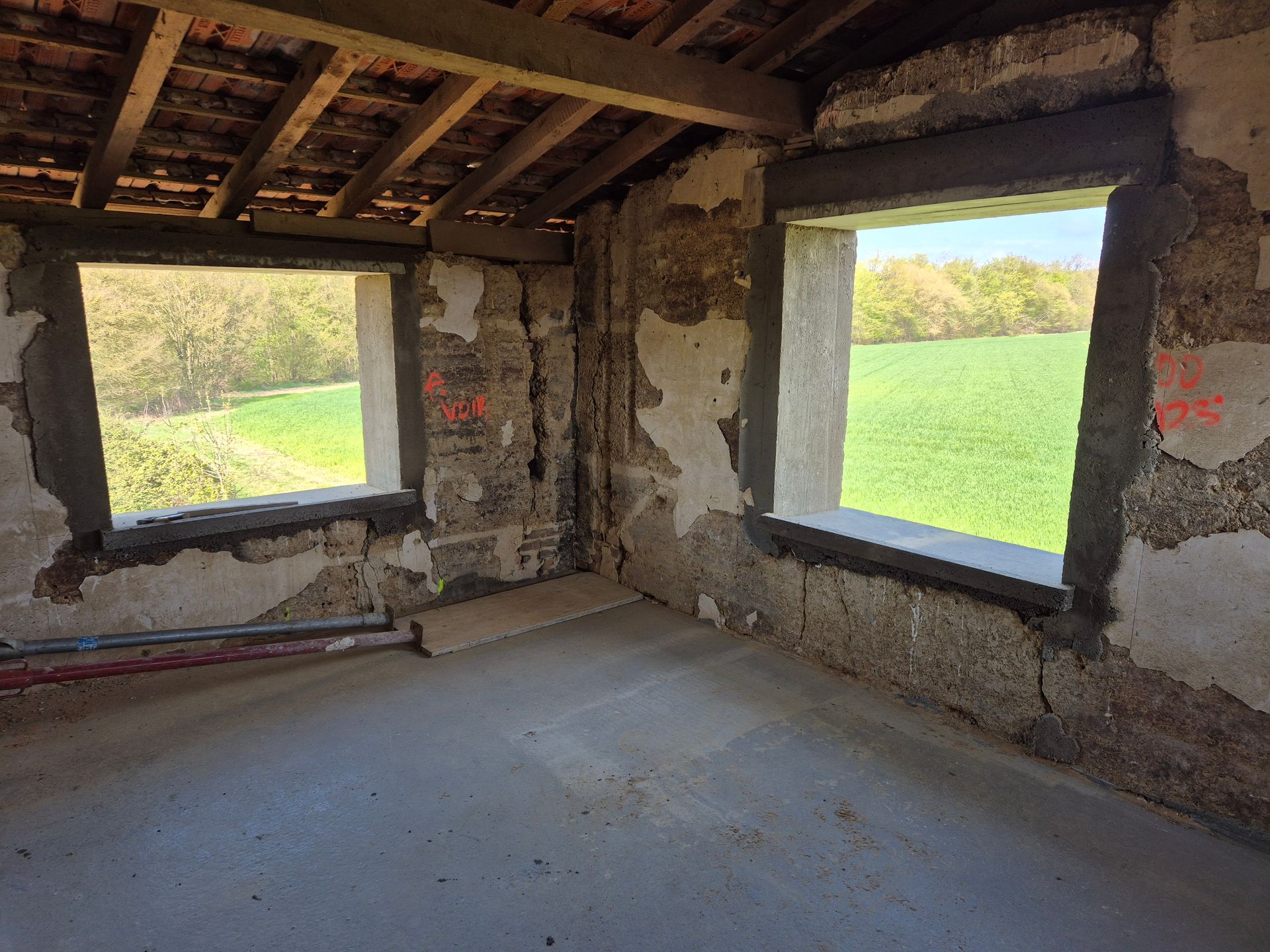 Intérieur d'une pièce endommagée et inachevée, avec poutres apparentes, murs bruts et deux fenêtres ouvertes
