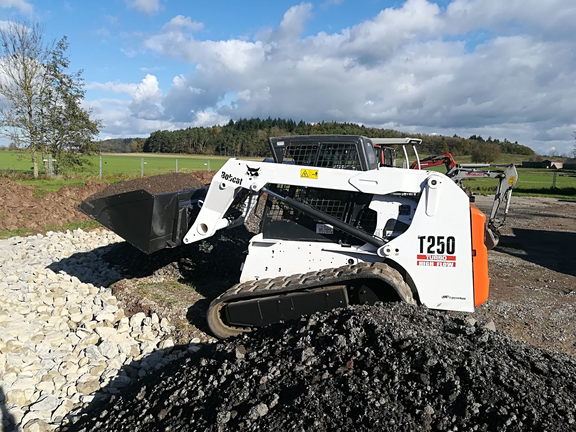 Weingärtner Baumaschinen – Kompaktlader Bobcat T250 zwischen mehreren Erd- und Gesteinshaufen
