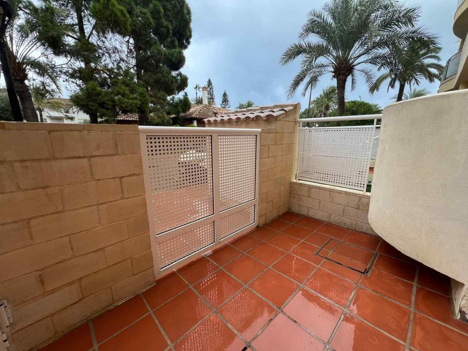 Patio exterior con suelo de baldosas rojas, paredes beige y una verja blanca de celosía bajo las palmeras.