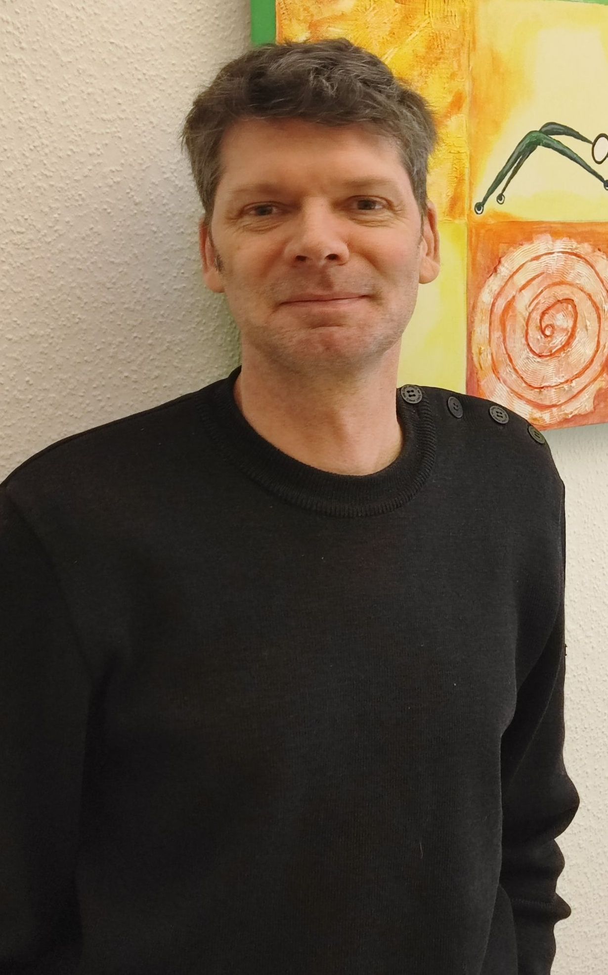 Portrait Thomas Junker - Ein Mann in einem schwarzen Pullover steht vor einem Gemälde an einer Wand.
