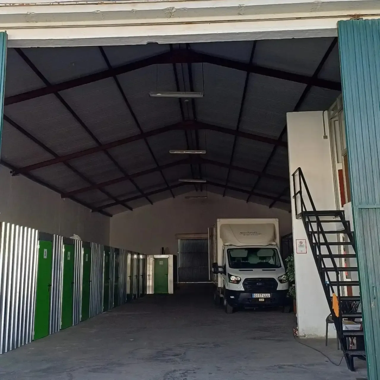 Interior de un almacén. Puertas verdes y taquillas metálicas bordean las paredes, con una furgoneta estacionada en el centro.