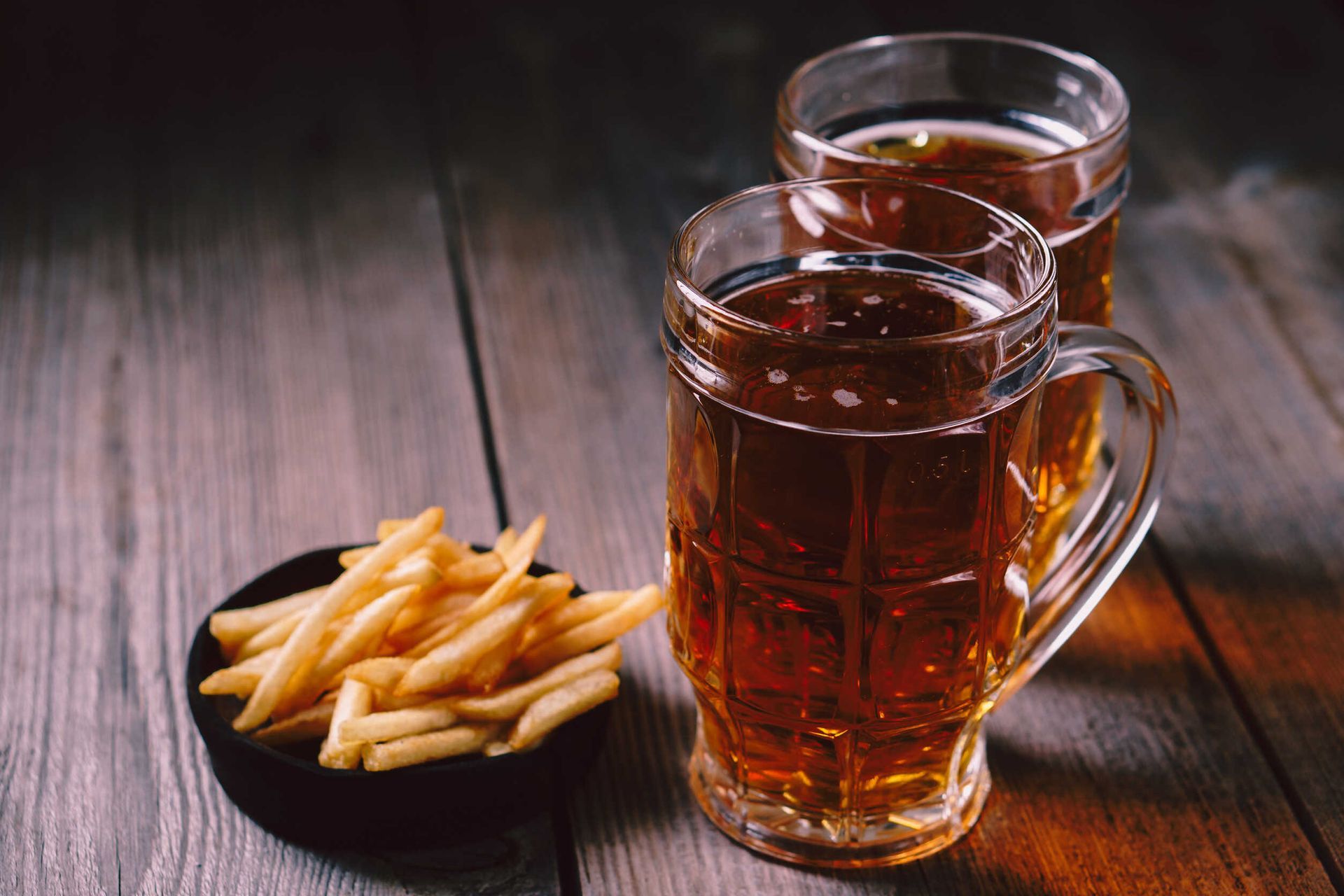Dos jarras de cerveza y un bol de patatas fritas en una mesa de madera.