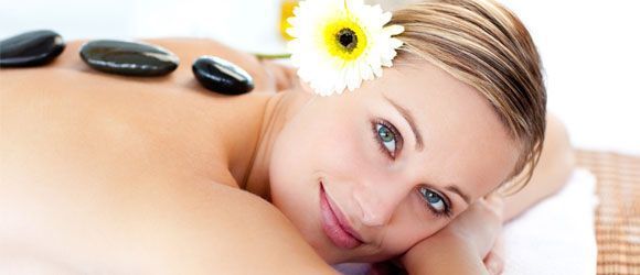 Mujer recibiendo masaje con piedras calientes, con flor en el cabello, sonriendo, relajándose en una mesa de masajes.