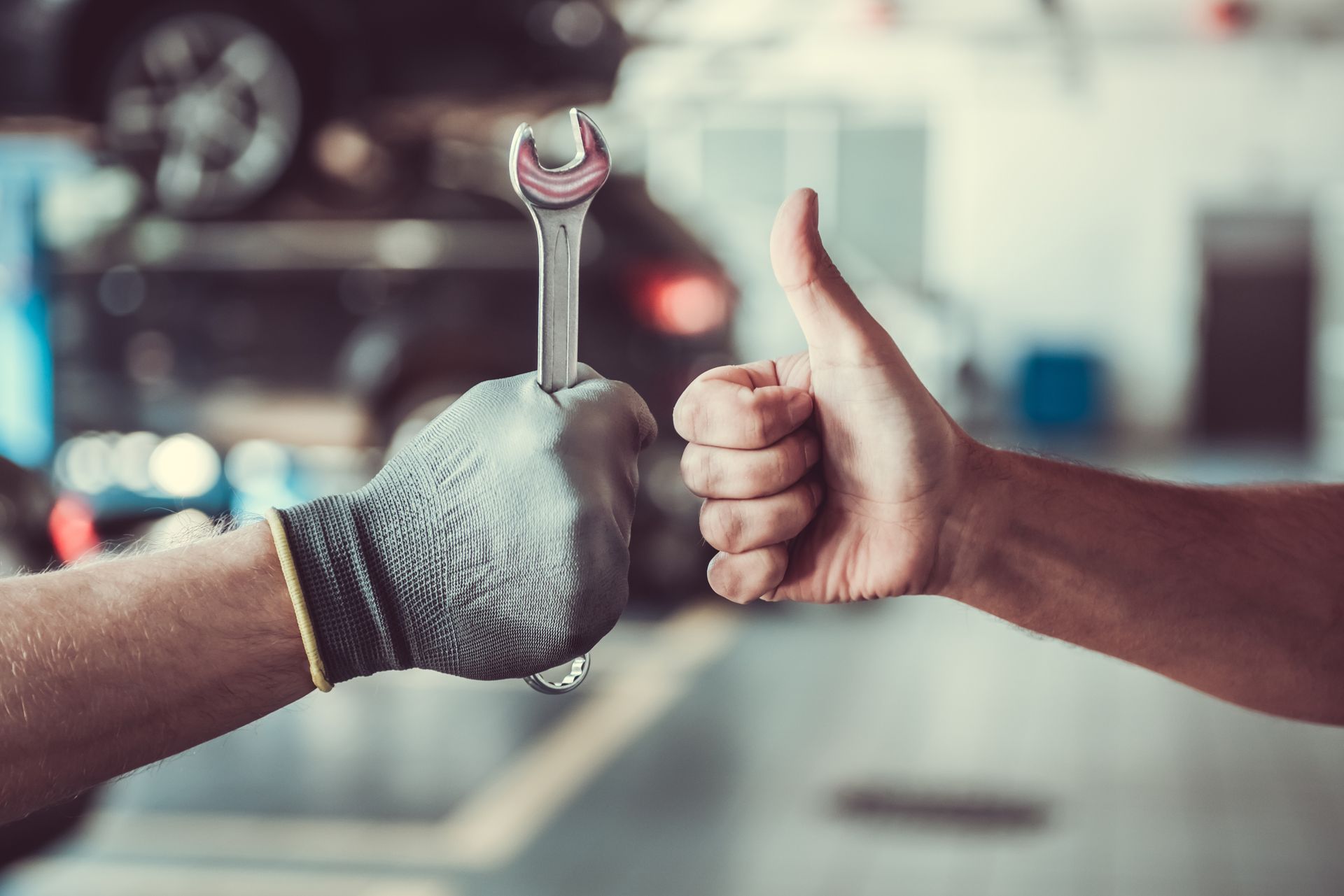 Des mains faisant un pouce levé et tenant une clé à molette dans un atelier de réparation automobile.