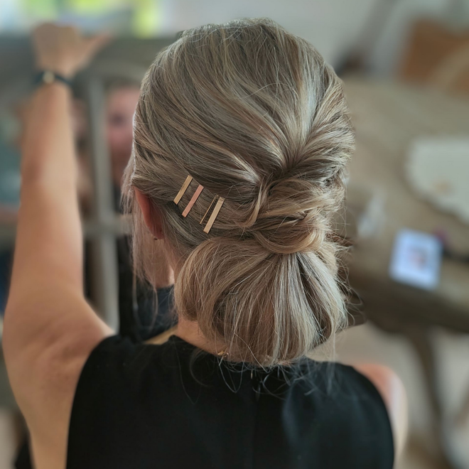 Femme aux cheveux châtain clair relevés et ornés d'épingles dorées. Elle porte un haut noir dans une pièce lumineuse.