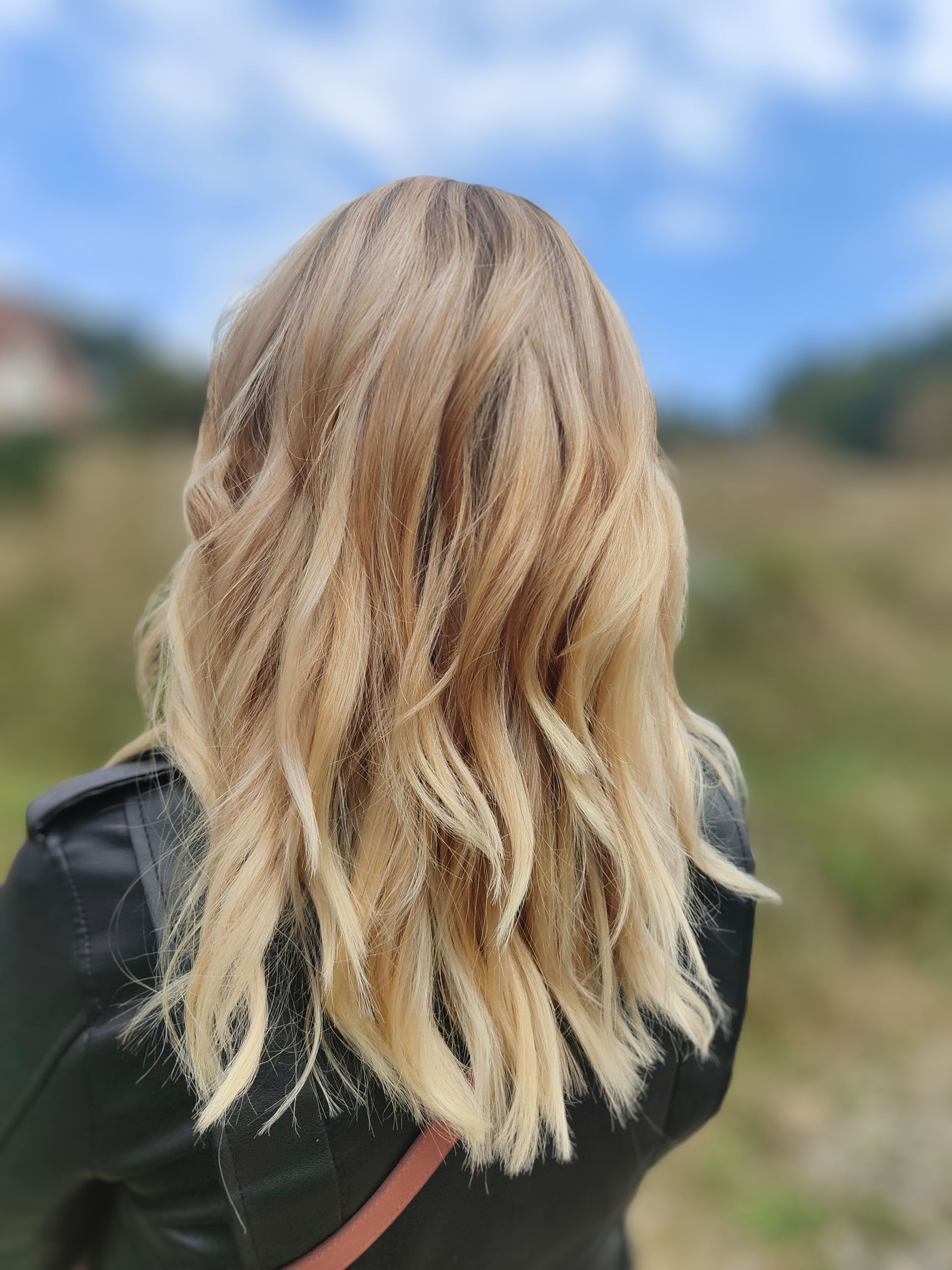 Cheveux blonds, légèrement ondulés, sur une personne portant une veste noire, à l'extérieur avec un arrière-plan flou.