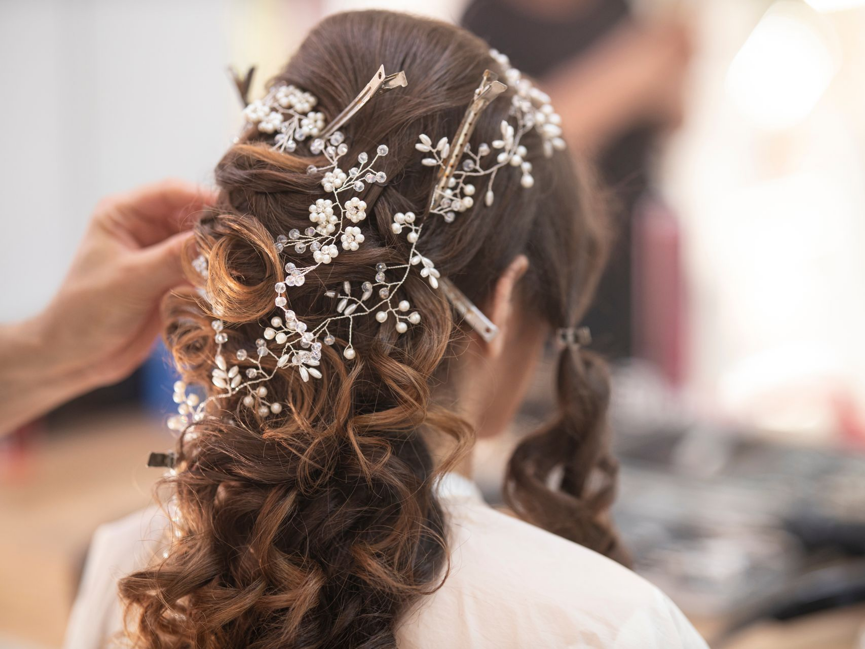 Personne se faisant coiffer ; postiche floral blanc orné ; mains du styliste ajustant le chignon.