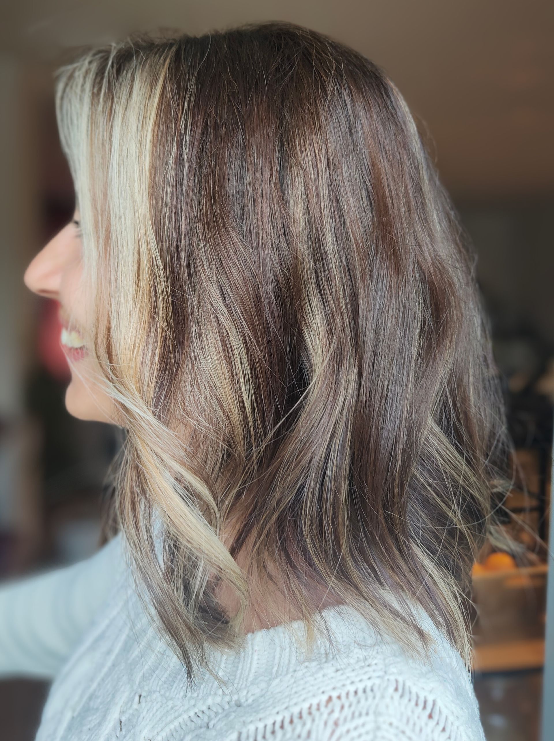 Femme aux cheveux bruns et blonds frisés, souriant devant une fenêtre.