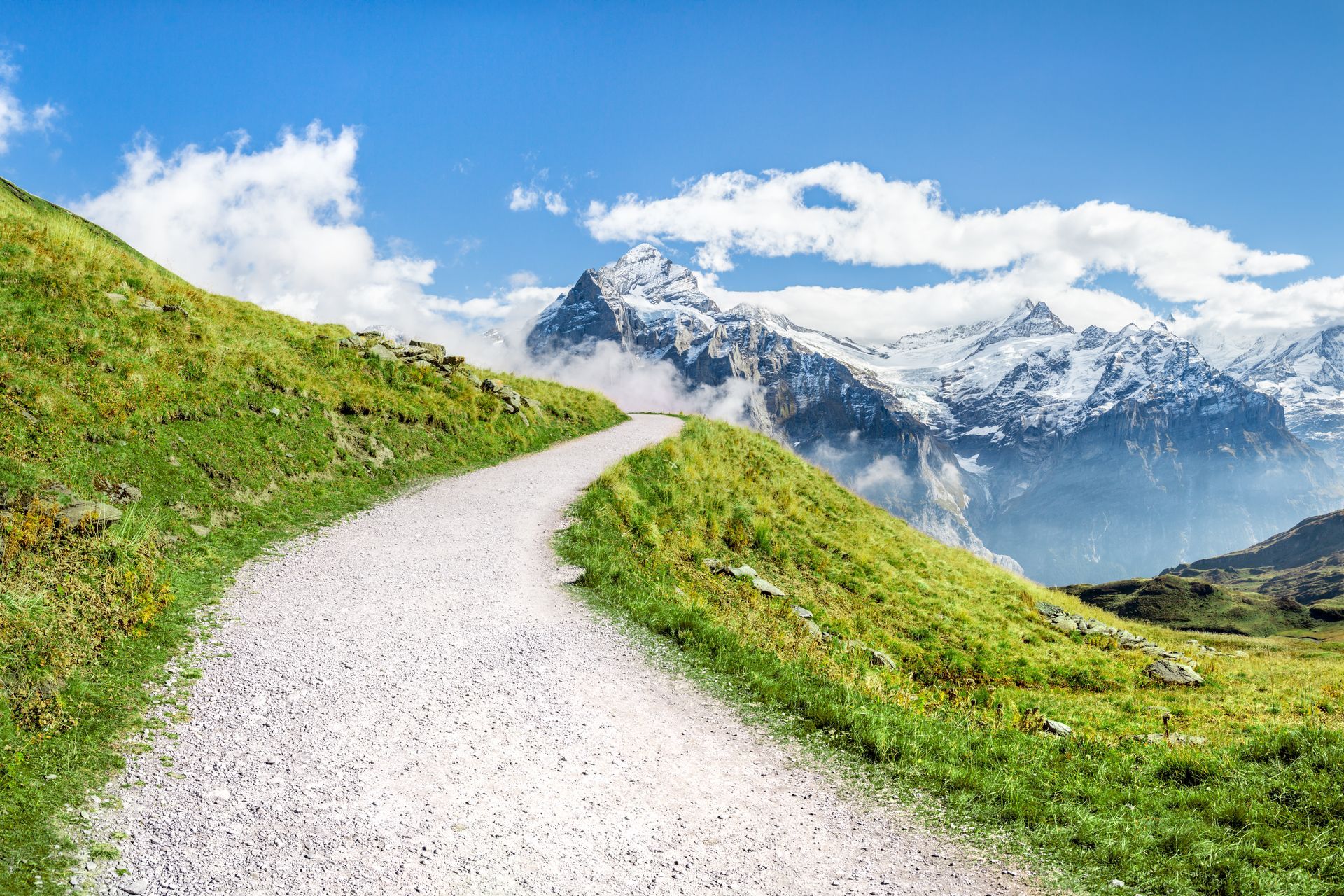 Ein gewundener Schotterweg an einem grasbewachsenen Hang führt zu schneebedeckten Bergen unter einem blauen Himmel mit Wolken.