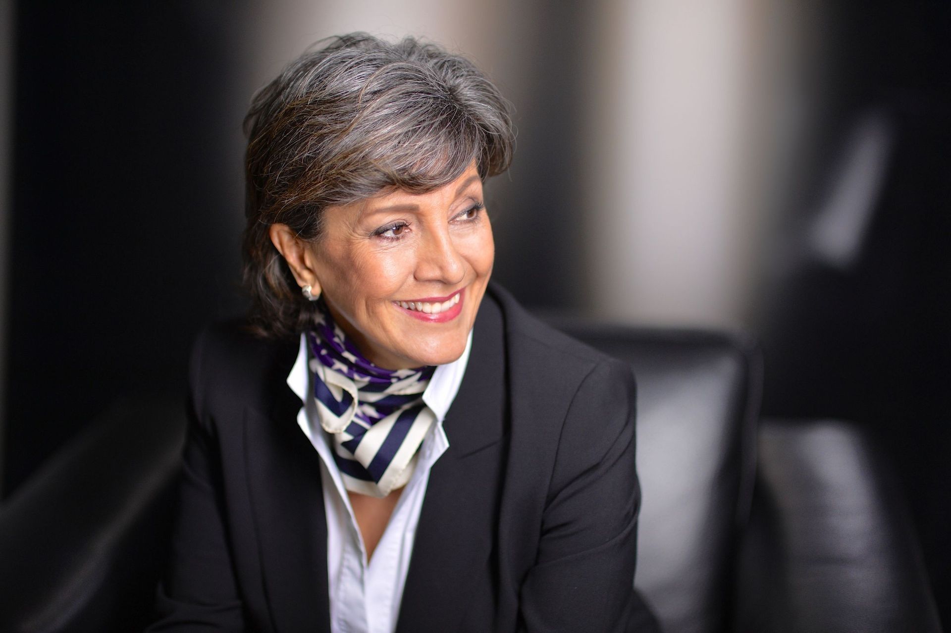 Frau in schwarzem Blazer lächelt, weisses Hemd, gemusterter Schal. Dunkler Hintergrund, professionelles Umfeld.