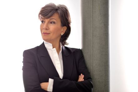 Frau in schwarzem Anzug, die Arme verschränkt, lehnt an einer grauen Säule und blickt zur Seite.