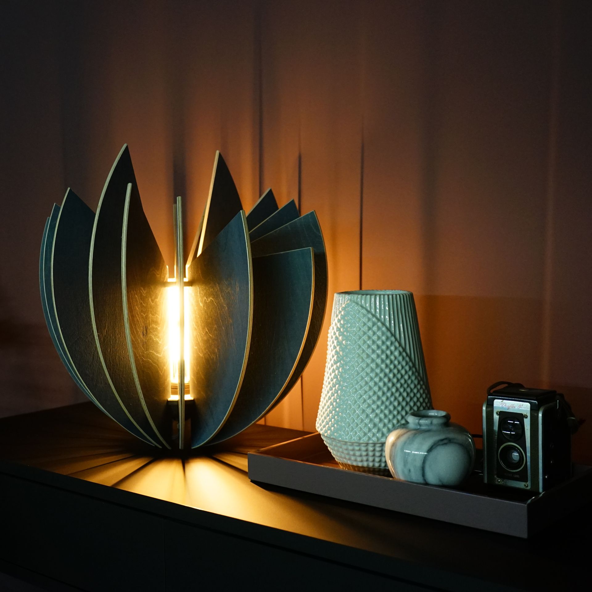 Eine beleuchtete, moderne Lampe in Blütenblattform steht auf einem Tisch neben Vasen und einer Kamera und spendet warmes Licht.