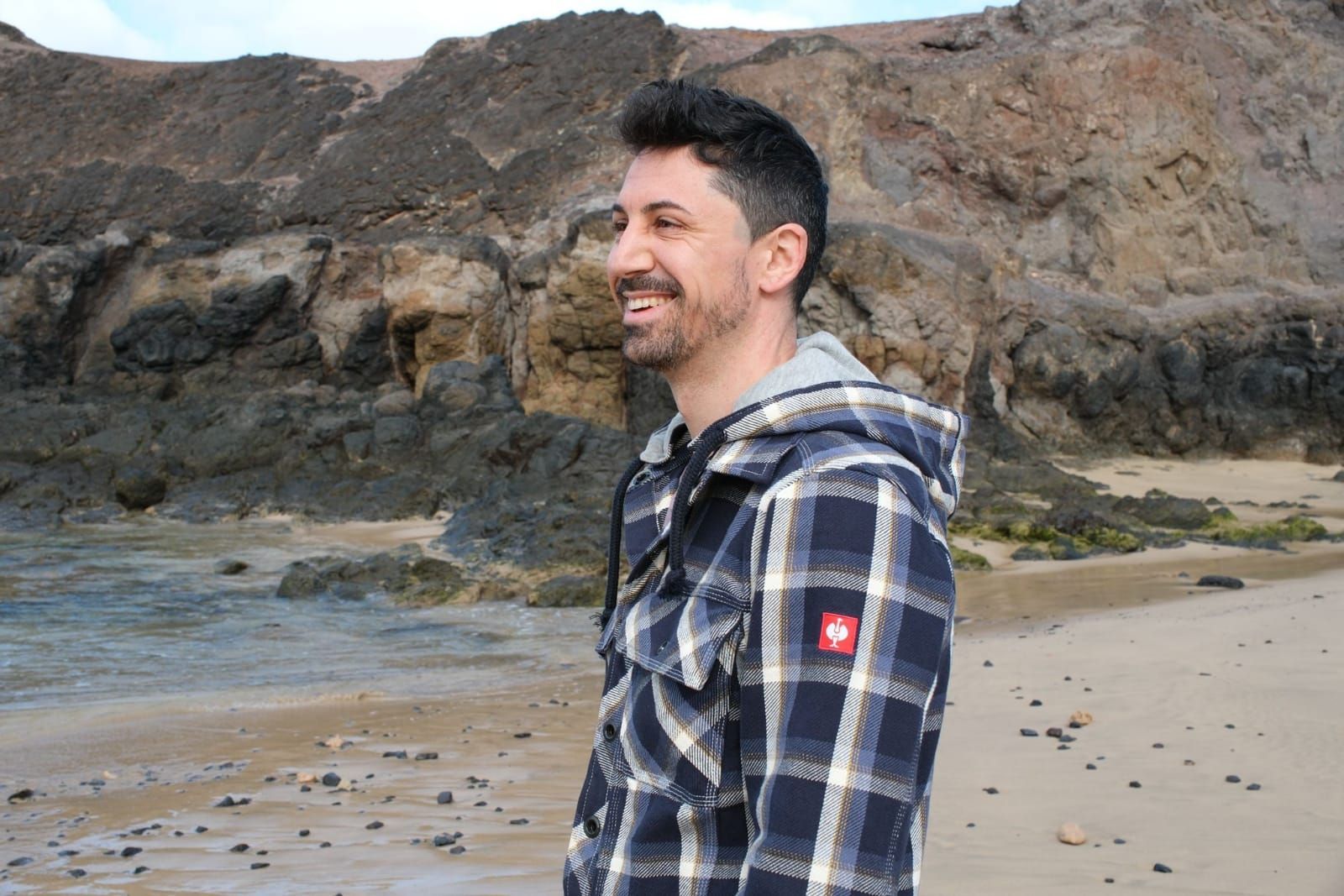 Ein Mann mit Sonnenbrille, weissem Hemd und blauen Shorts steht an einem sonnigen Tag an einer Steinmauer mit Blick auf den Ozean.