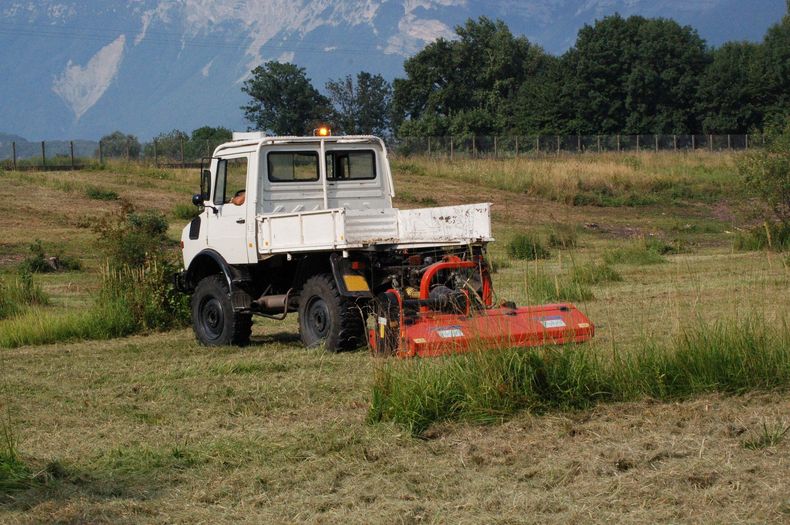 Un camion utilitaire blanc tond l'herbe dans un champ avec des montagnes en arrière-plan.