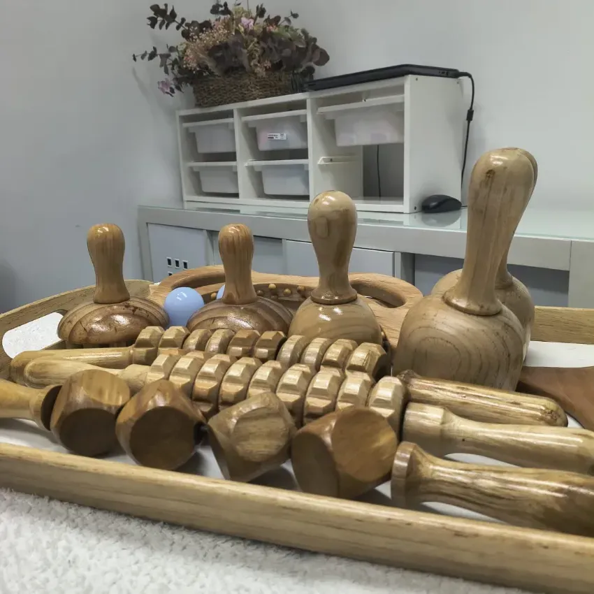 En una habitación blanca con estanterías y flores secas, se disponen herramientas de masaje de madera sobre una bandeja.