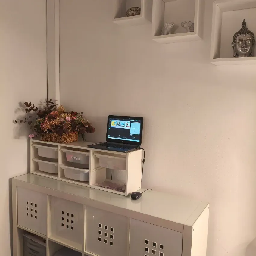 Un pequeño escritorio con un ordenador portátil y cajones contra una pared blanca, con estantes y una estatua de Buda encima.