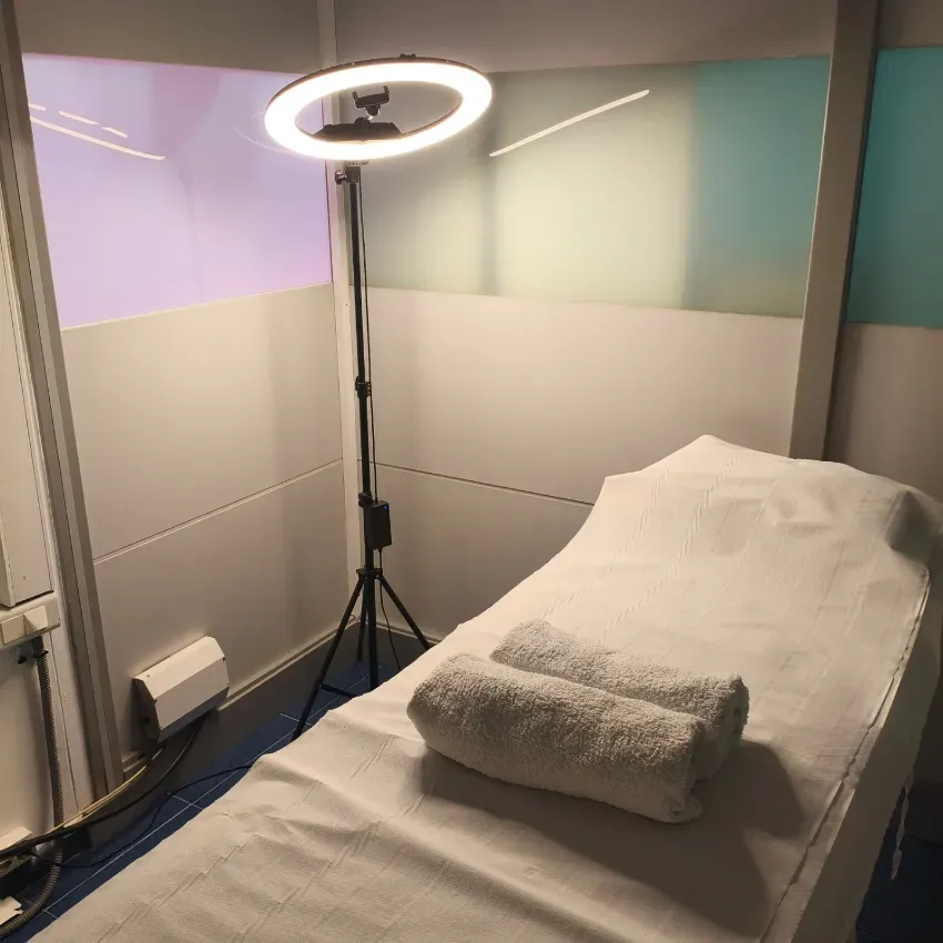 Sala de tratamiento compacta con camilla de masaje, toalla doblada y soporte con aro de luz iluminado.