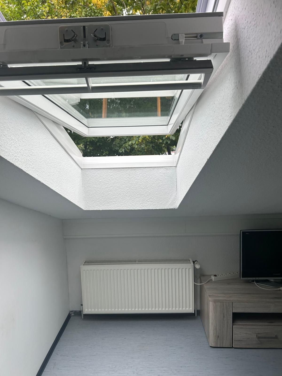 Kleines Dachzimmer mit Dachfenster, weißem Heizkörper und Schreibtisch mit Fernseher an der Wand.