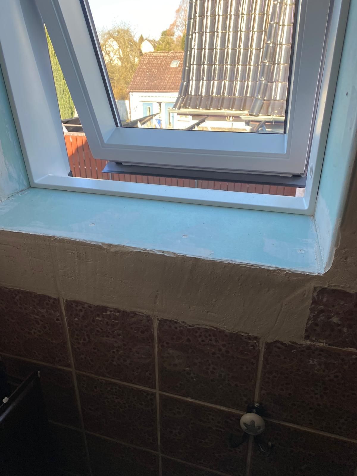 Ein offenes Dachfenster über einer gefliesten Badezimmerwand gibt den Blick auf die Dächer draußen frei.