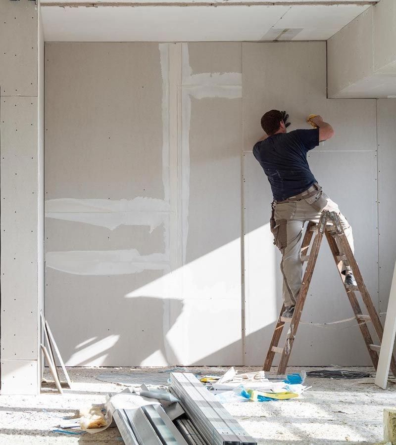 Un trabajador, subido a una escalera, repara el pladur de una habitación soleada en obras.