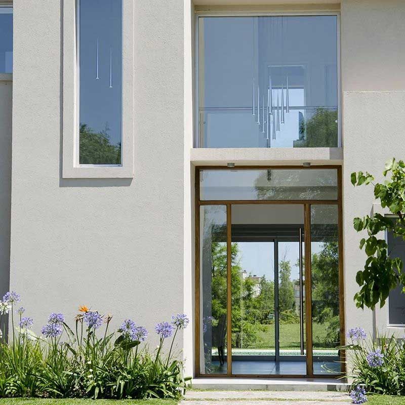 Fachada de casa moderna con grandes ventanales, puerta de cristal y flores moradas en un jardín.