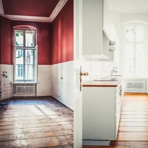 Interior de una habitación vacía con paredes rojas y blancas, una ventana alta y suelo de madera, junto a una cocina blanca y luminosa.