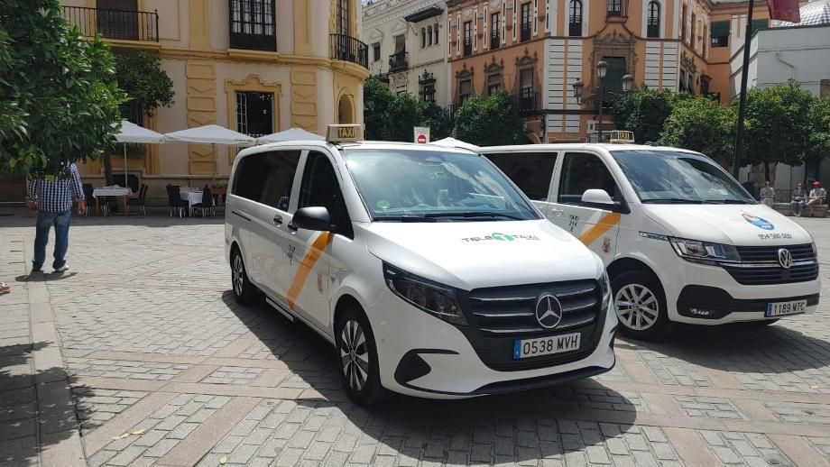 Dos taxis blancos estacionados en una plaza adoquinada frente a edificios.
