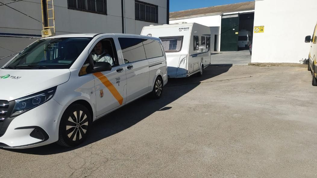 Una furgoneta blanca con una caravana acoplada circula por una zona pavimentada, bajo la luz del sol.