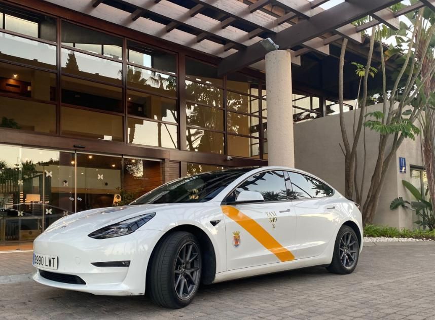 Taxi Tesla blanco estacionado afuera de un edificio con puertas de vidrio y franja amarilla en el costado.