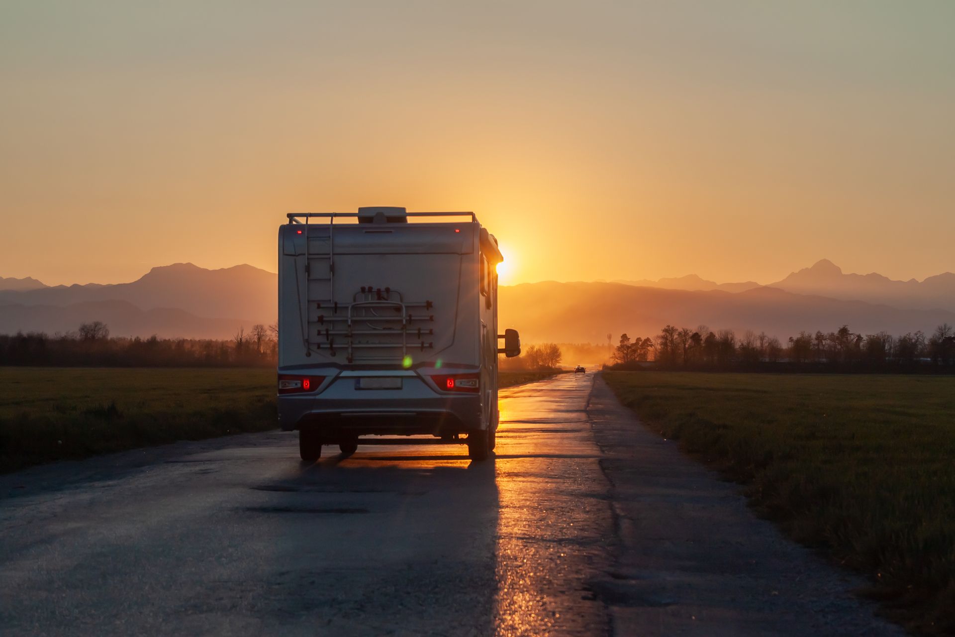 Mit dem Wohnmobil fährt man bei Sonnenuntergang eine nasse Straße entlang, die Sonne steht hinter den Bergen, Felder erstrecken sich zu beiden Seiten.