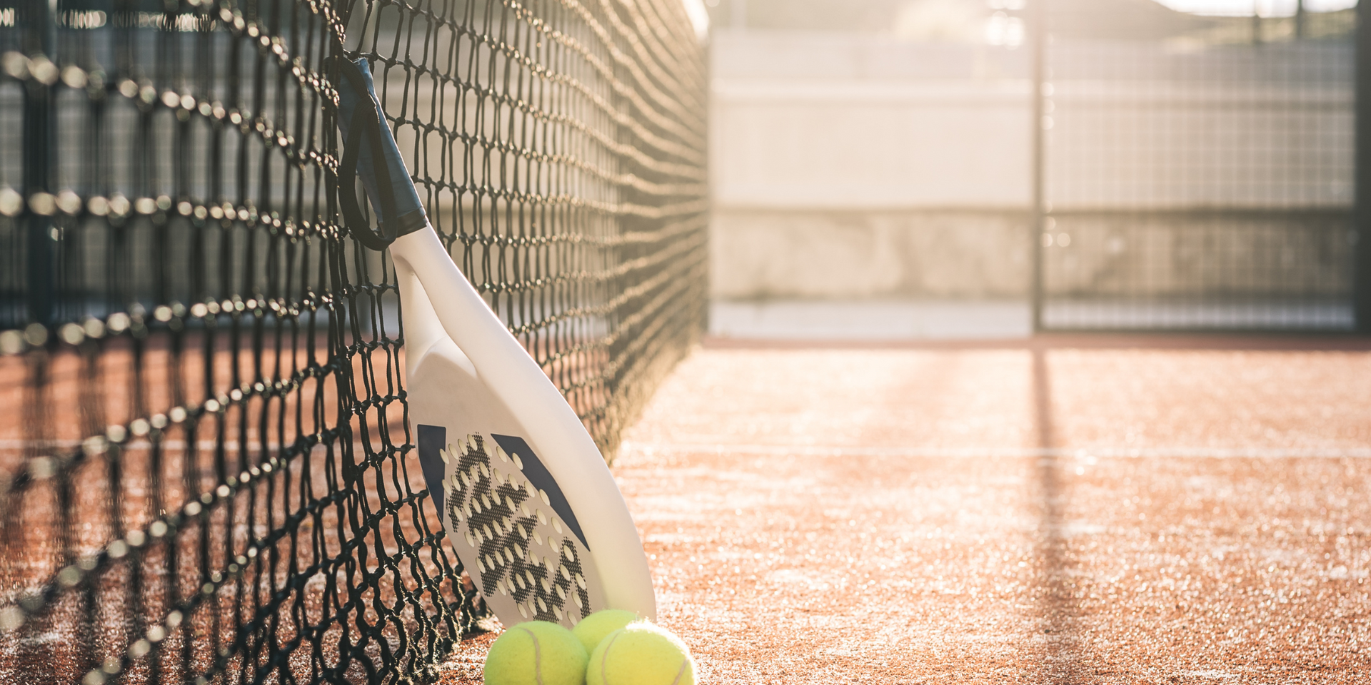Una raqueta de tenis y tres pelotas de tenis están apoyadas contra una red de tenis en una cancha de tenis.