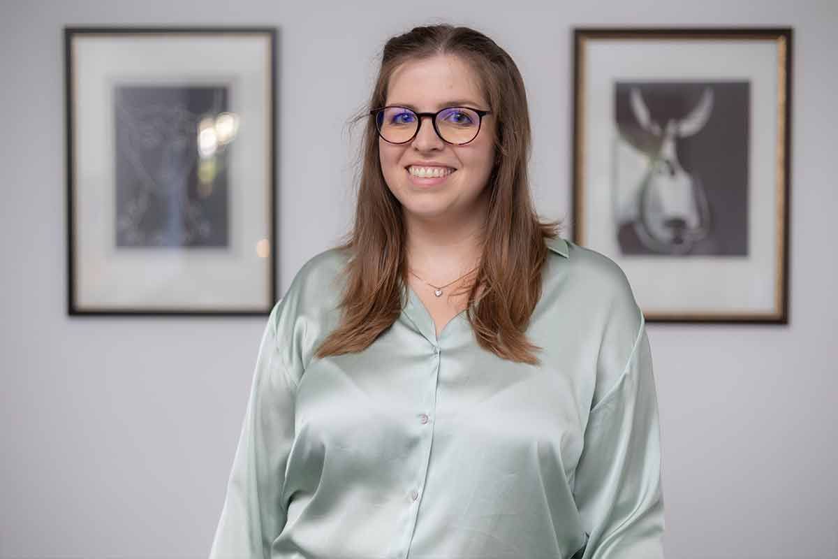 Eine Frau mit Brille und grünem Hemd steht vor einer Wand mit Bildern daran.