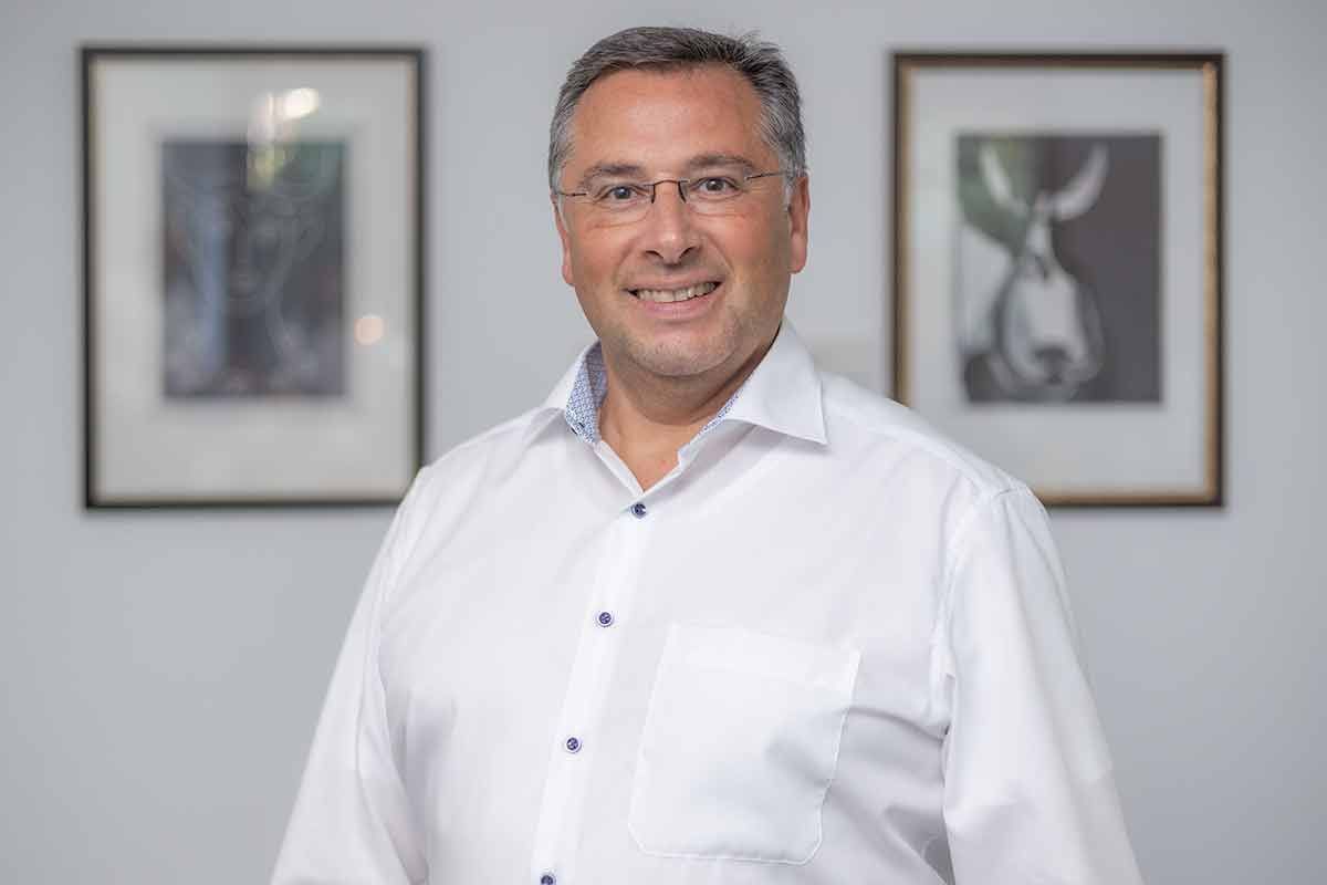 Ein Mann in einem weißen Hemd und mit Brille steht vor zwei gerahmten Bildern.