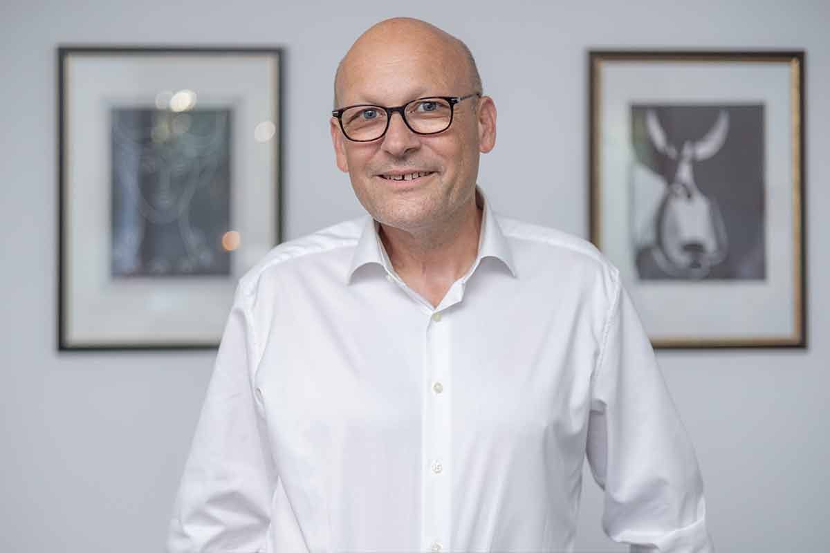 Ein kahlköpfiger Mann mit Brille und weißem Hemd steht vor zwei gerahmten Bildern.